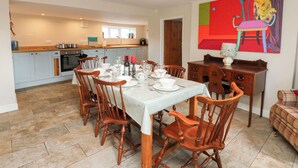 A dining area with a table set for four, wooden chairs, a kitchen in the background, and a colorful abstract painting on the wall. - Rosemary Cottage (Leyburn)