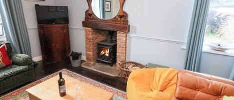 Un salon chaleureux avec une cheminée en briques, une table basse en bois sur laquelle se trouvent une bouteille et un verre, un canapé vert et un fauteuil poire orange.