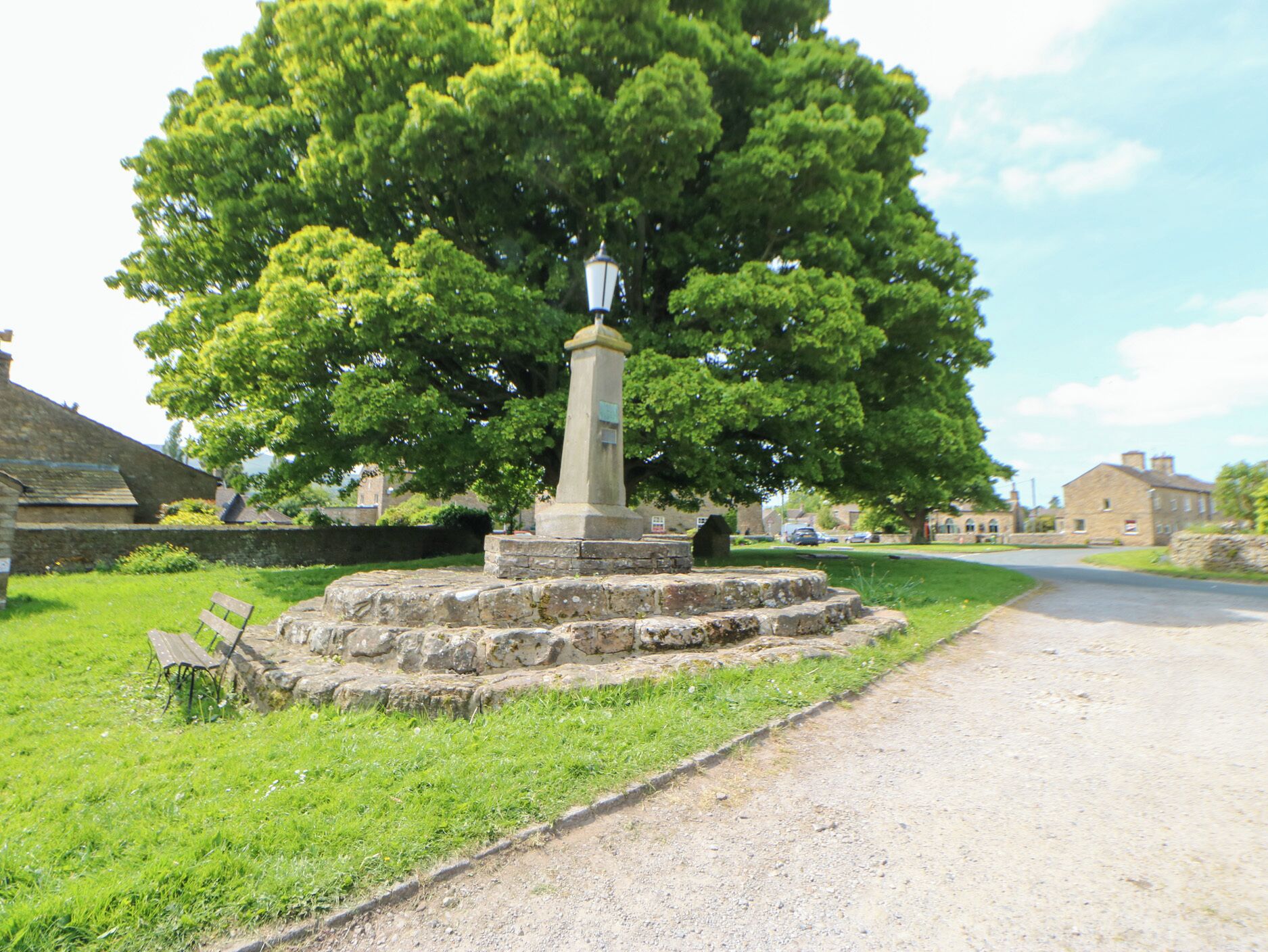 A stone monument with a statue, surrounded by grass and a tree, located at a roadside intersection.