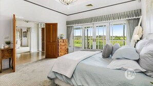 A spacious bedroom with a large bed, a wooden dresser, and a view of a green landscape through the window.