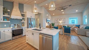 A modern kitchen with a central island, stainless steel appliances, and a dining area with a sofa and decorative pillows. - Pool, Firepit + Golf Cart Zone + Near Beach Looney Dunes by Avantstay (Port Aransas)