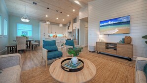 A spacious living area with a kitchen in the background, a large TV mounted on the wall, and a dining area with wicker furniture. - Pool, Firepit + Golf Cart Zone + Near Beach Looney Dunes by Avantstay (Port Aransas)