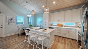 A modern kitchen with white cabinetry, a central island with bar stools, pendant lighting, and a view of the outdoors through the windows. - Pool, Firepit + Golf Cart Zone + Near Beach Looney Dunes by Avantstay (Port Aransas)
