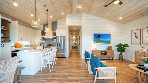 A spacious kitchen with a central island, a dining area with a view, and a living room with a beach-themed TV. - Pool, Firepit + Golf Cart Zone + Near Beach Looney Dunes by Avantstay (Port Aransas)