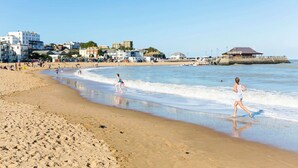 Una playa con gente caminando por la orilla, edificios al fondo y un muelle que se adentra en el agua.
