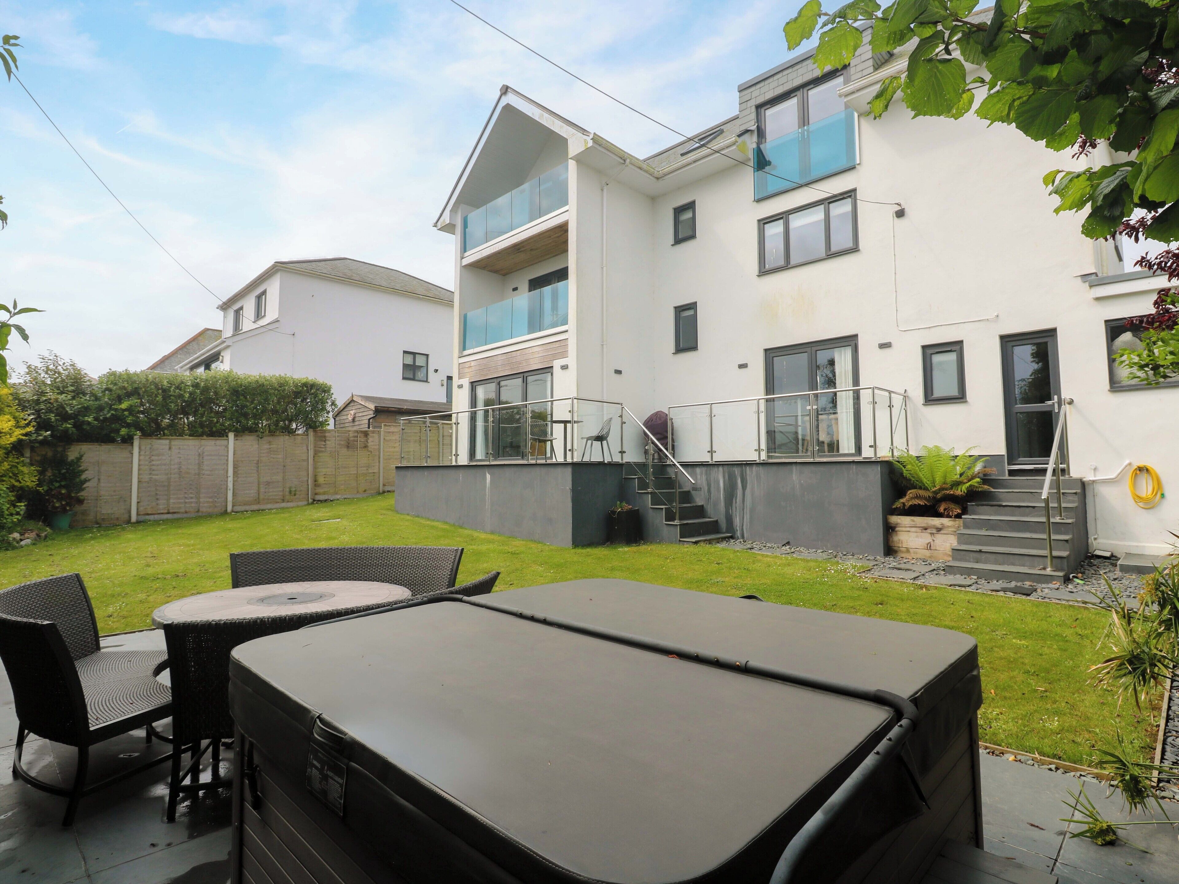 A modern two-story house with a balcony, a patio with outdoor furniture, and a well-maintained garden.