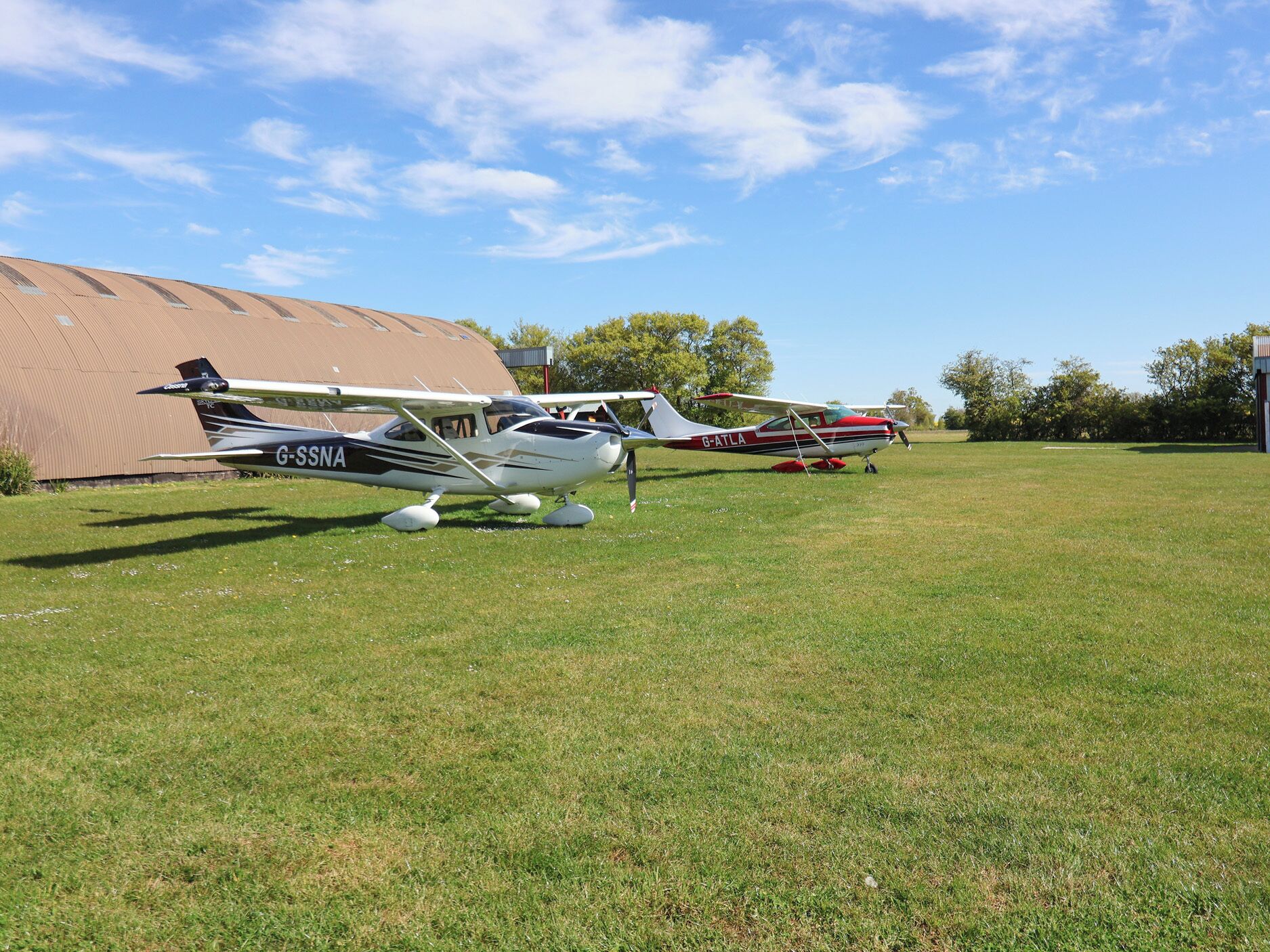 Two small aircraft parked on a grassy field with a hangar in the background.