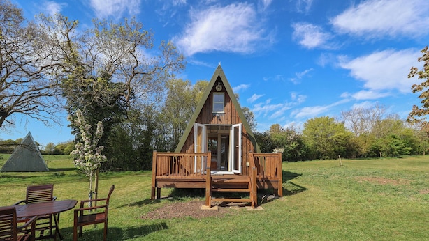 A wooden cabin with a triangular roof, a deck, and a table with chairs. - A-frame (Woodbridge)