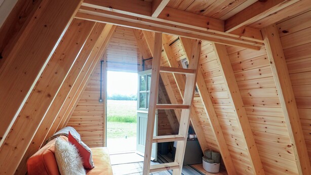 A wooden attic bedroom with a loft bed, a ladder, and a view of a green field. - Robin (Woodbridge)