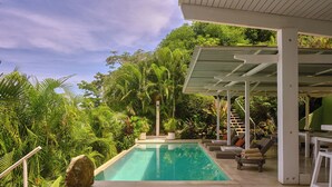 A pool with a covered area, surrounded by lush greenery and a staircase leading to a roofed section.