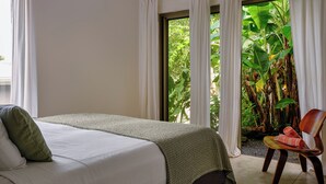 A bedroom with a bed, a chair, and a view of lush greenery through the window.
