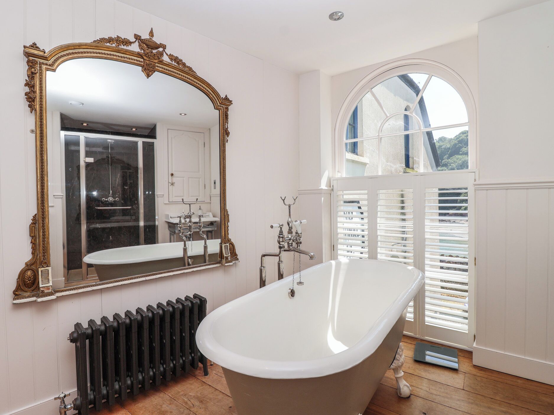 Un baño con una bañera independiente, un espejo grande y ornamentado, y un ventanal con contraventanas blancas.