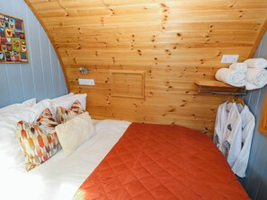 A room with a wooden ceiling, a bed with white and orange bedding, a wall-mounted shelf with towels, and a colorful artwork on the wall. - Ty Twt Teifi (Tregaron)