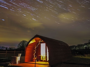 A wooden structure with an open archway, illuminated by warm interior lighting, set against a night sky with visible star trails. - Ty Twt Teifi (Tregaron)