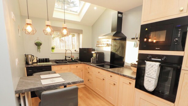 A modern kitchen with a dining area, featuring a built-in oven, microwave, and a large window providing natural light. - 10 Keekle Terrace (Cleator Moor)
