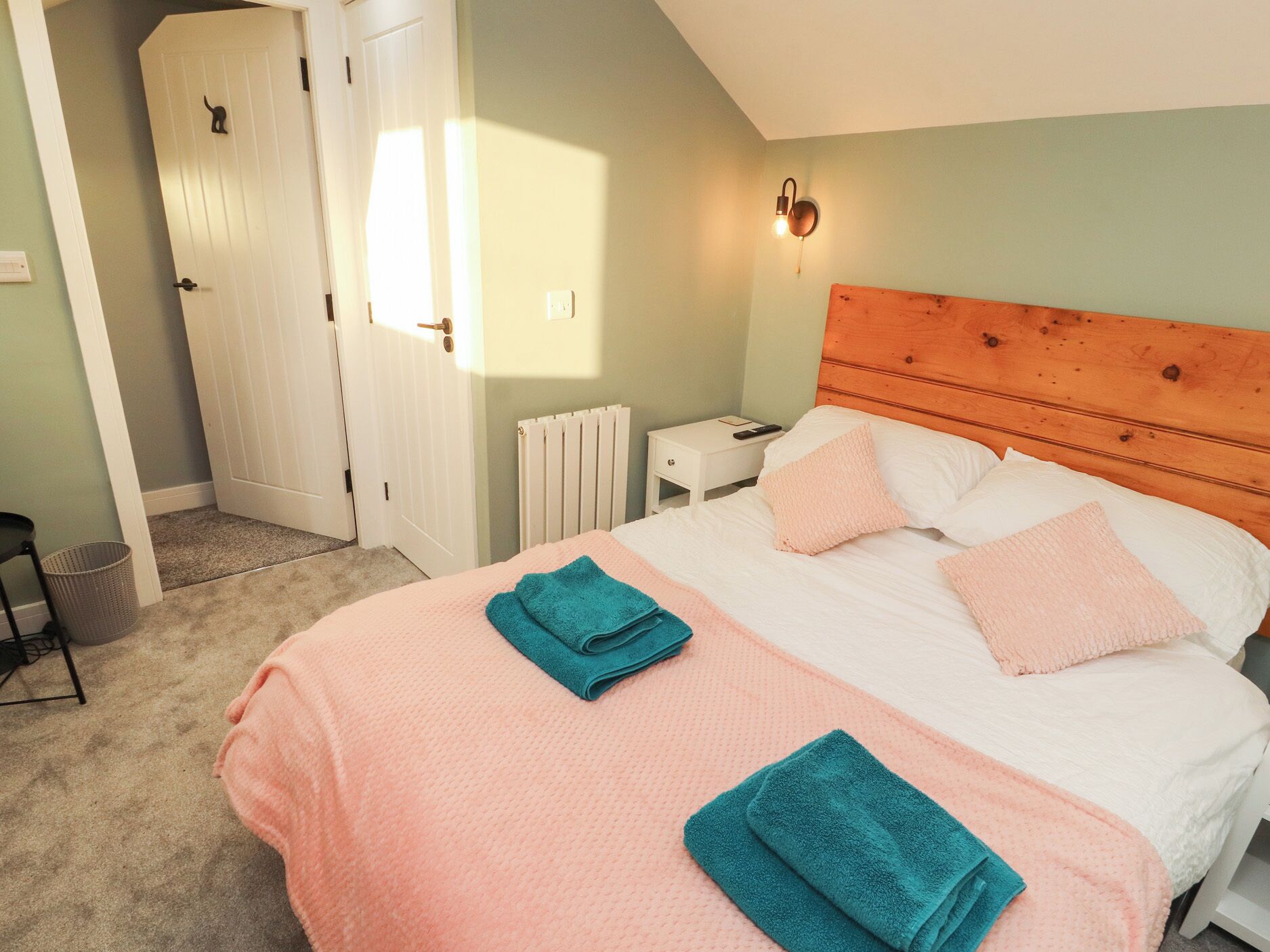 Un dormitorio con cabecera de madera, mesita de noche blanca, y una colcha rosa con almohadas en tonos rosa y verde azulado.