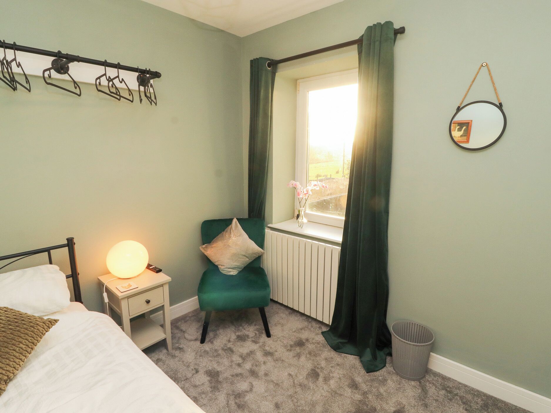 Un dormitorio con cama, mesita de noche, silla, ventana con cortinas, radiador y espejo montado en la pared.