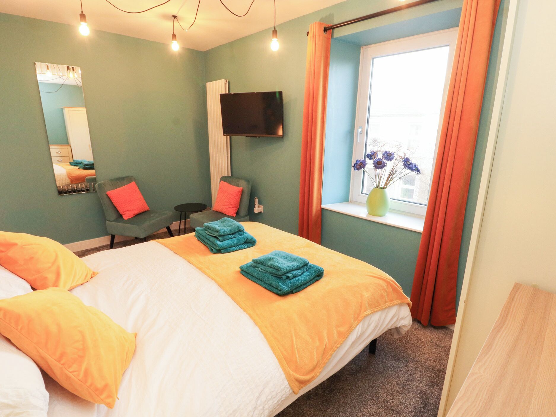 Habitación de hotel con cama, dos sillones, televisor, ventana con cortinas y un jarrón con flores.
