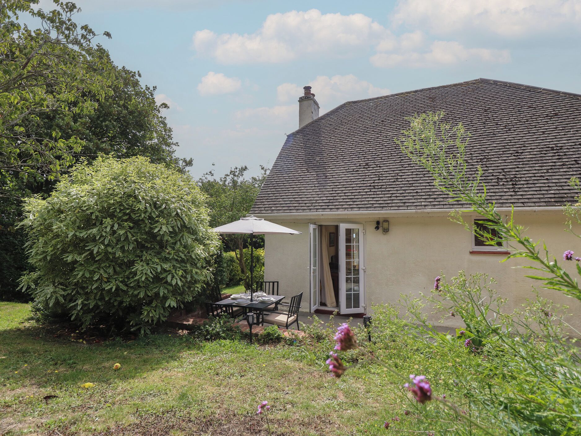 Ein Haus mit überdachter Terrasse, Schornstein und Garten mit Tisch und Stühlen.