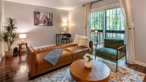 A living room with a brown leather sofa, a green armchair, a round wooden coffee table with a vase of flowers, a lamp, and a painting on the wall. - The Fern & Fable - King Bed/TownHome with Deck (Birmingham)