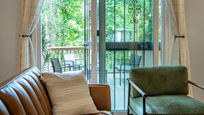 A room with a brown leather sofa and a green armchair, a glass door leading to an outdoor balcony with a table and chairs, and a view of greenery outside. - The Fern & Fable - King Bed/TownHome with Deck (Birmingham)