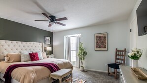 A bedroom with a tufted headboard, a large bed, a bench, a ceiling fan, a window with curtains, a potted plant, a wooden chair, a bedside table, and a framed picture on the wall.