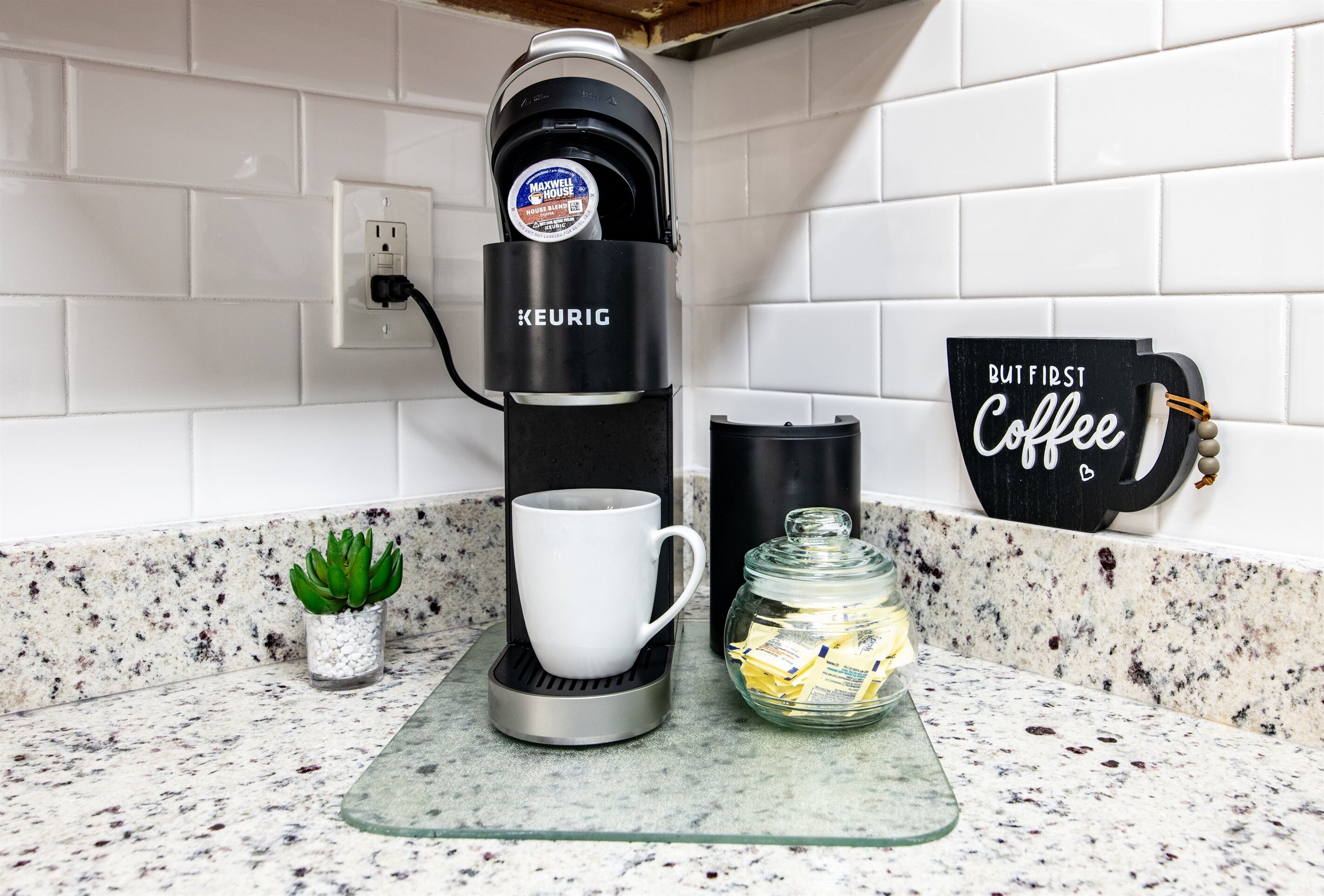 Une machine à café posée sur un plan de travail, avec une tasse, un bocal en verre et un panneau indiquant : « D’abord, un café ».