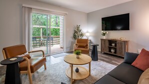 A modern living room with a sofa, armchair, and round coffee table, a flat-screen TV mounted on the wall, and a sliding glass door leading to an outdoor area.