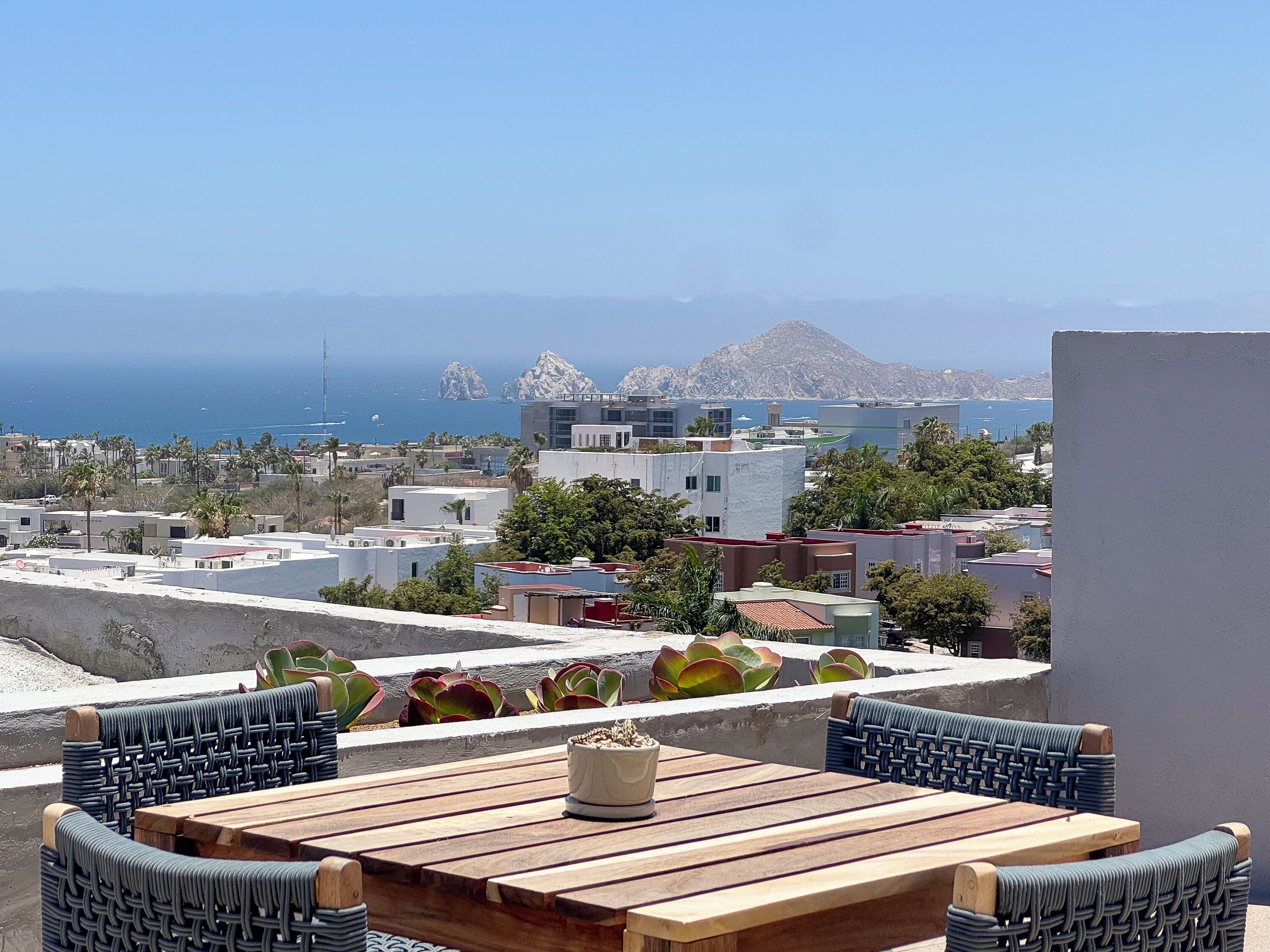 A terrace with a wooden table and wicker chairs, overlooking a coastal town with buildings and a mountain in the distance.
