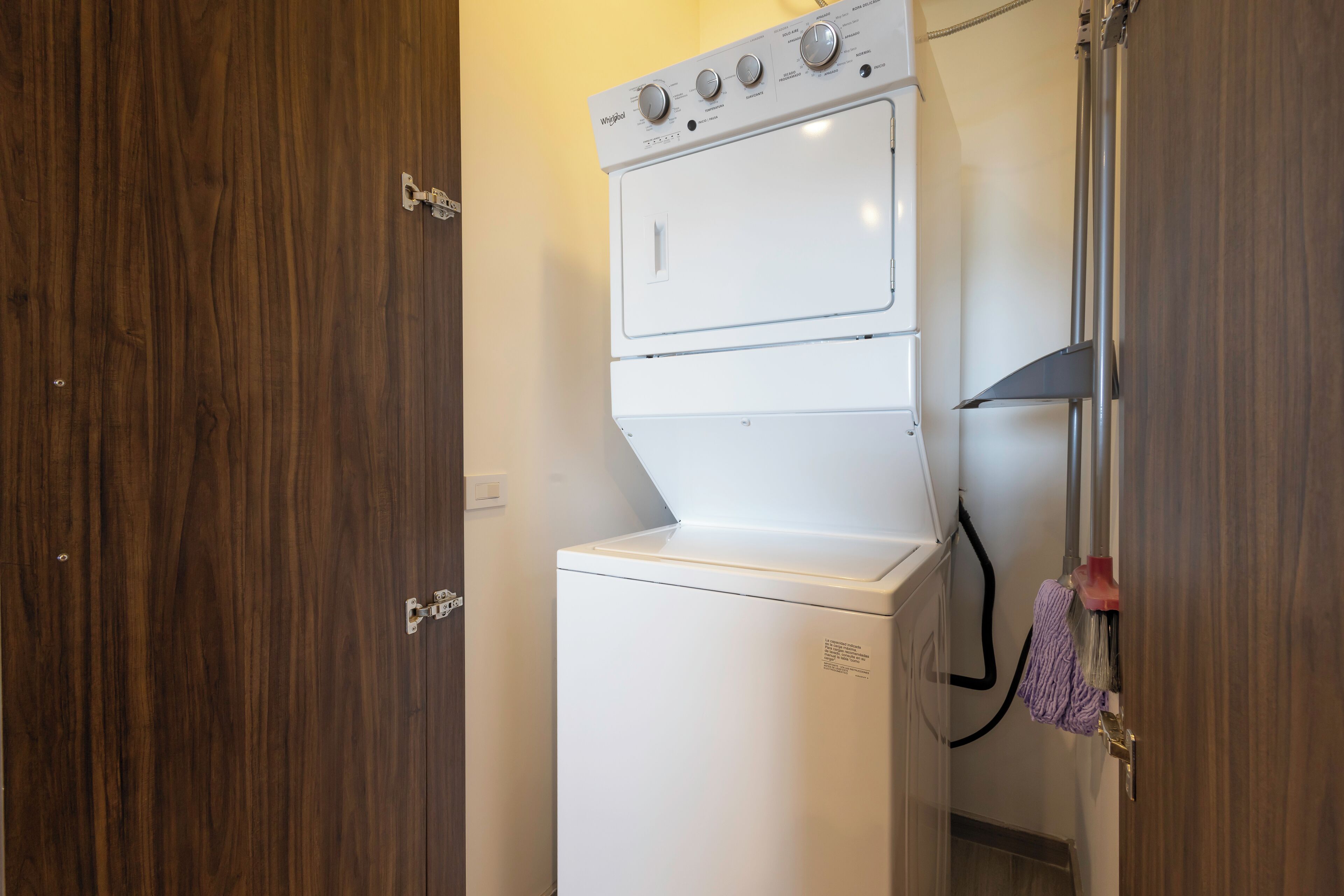 Een kleine, witte wasruimte met een ingebouwde wasmachine en droger, houten deuren en zichtbare leidingen.