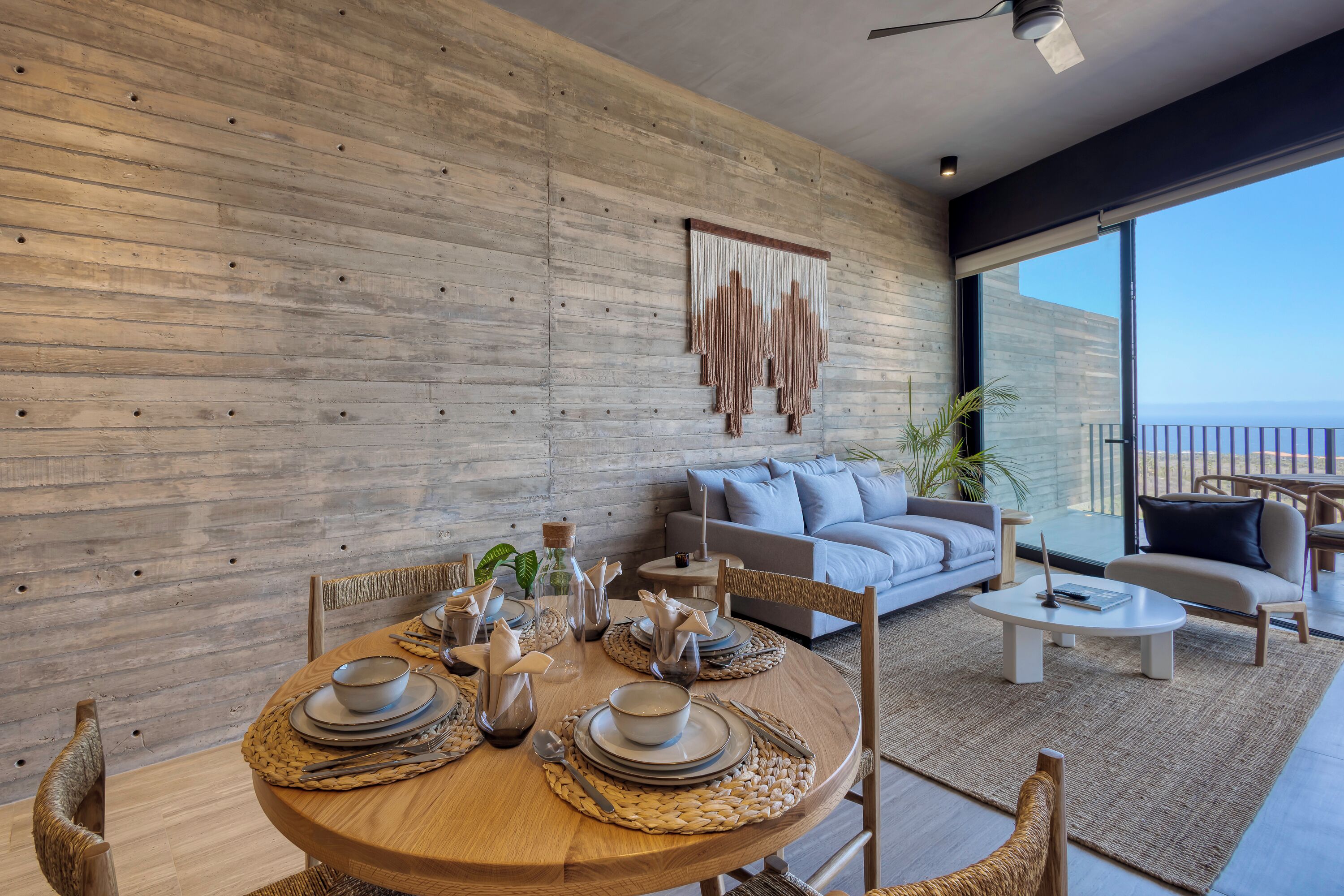 A modern dining area with a wooden table set for four, surrounded by wicker chairs. A large window offers a view of the ocean.