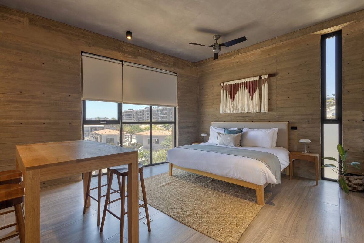 A modern bedroom with a large bed, a dining table, and a view of buildings through a sliding door.