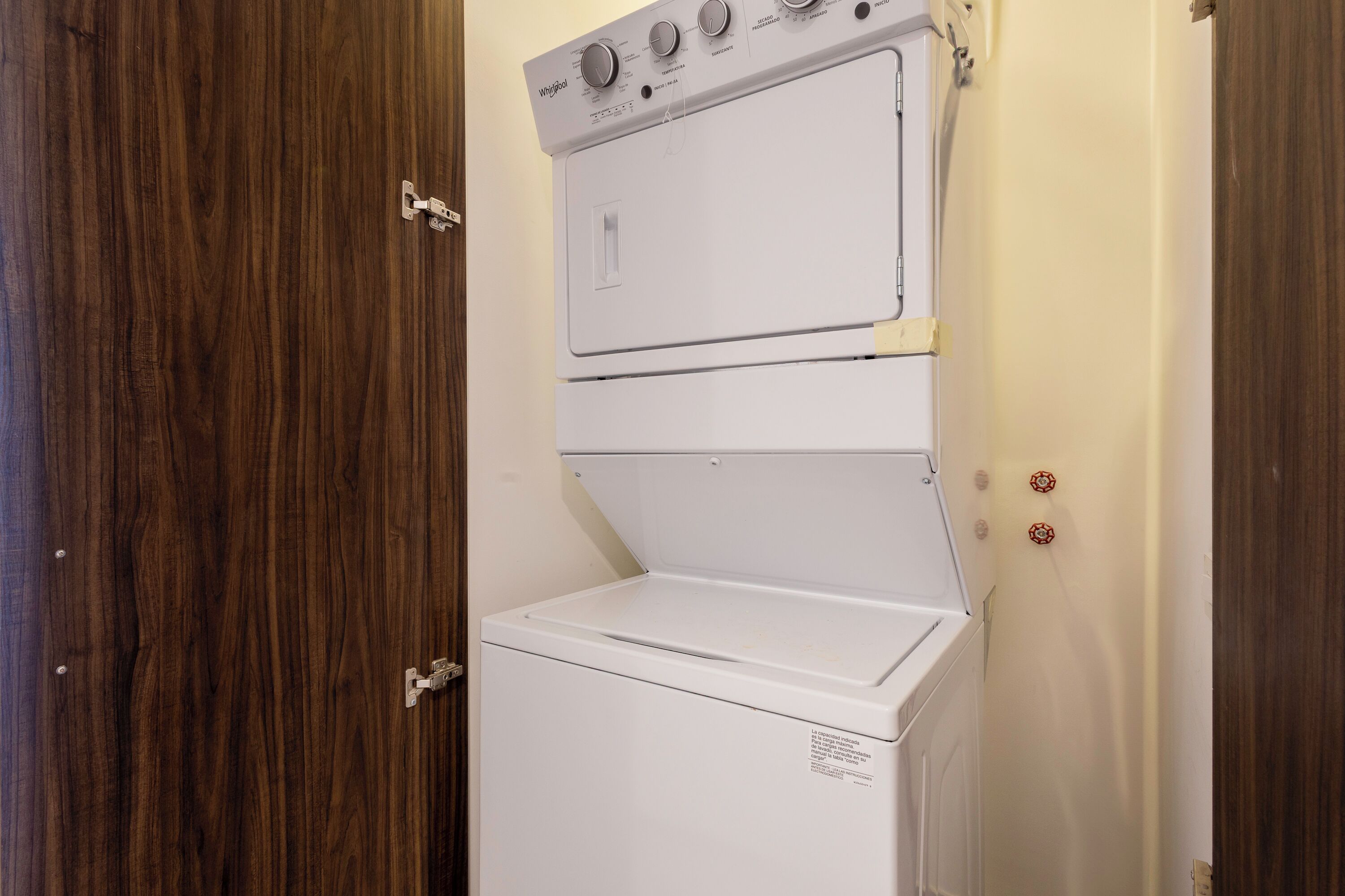 Una unidad compacta de lavadora y secadora apiladas, blanca, con un panel de control en la parte superior, instalada en una habitación con puertas de madera.