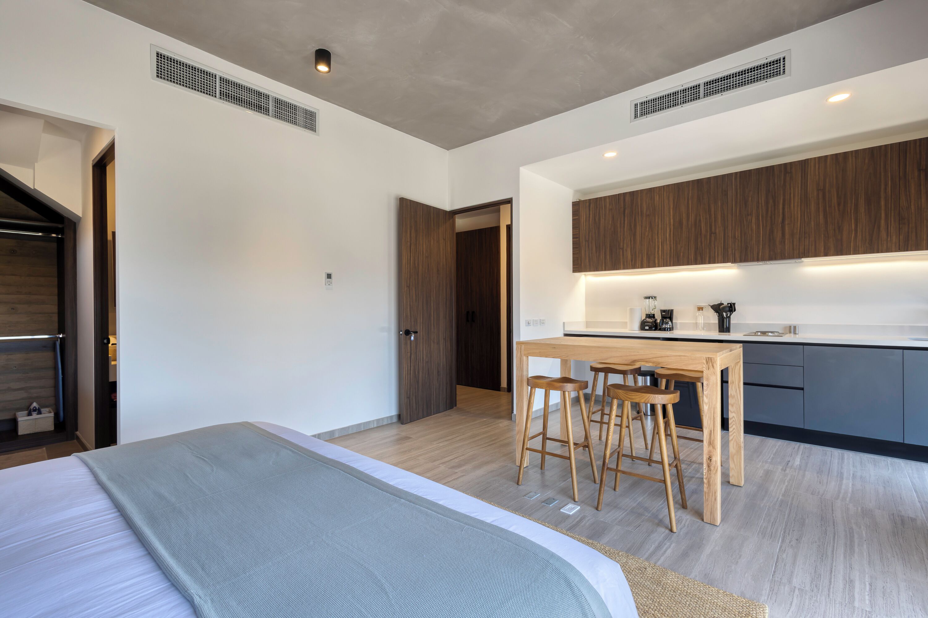 Een moderne slaapkamer met een groot bed, een houten eettafel met krukken, en een kitchenette met donkere kastjes.