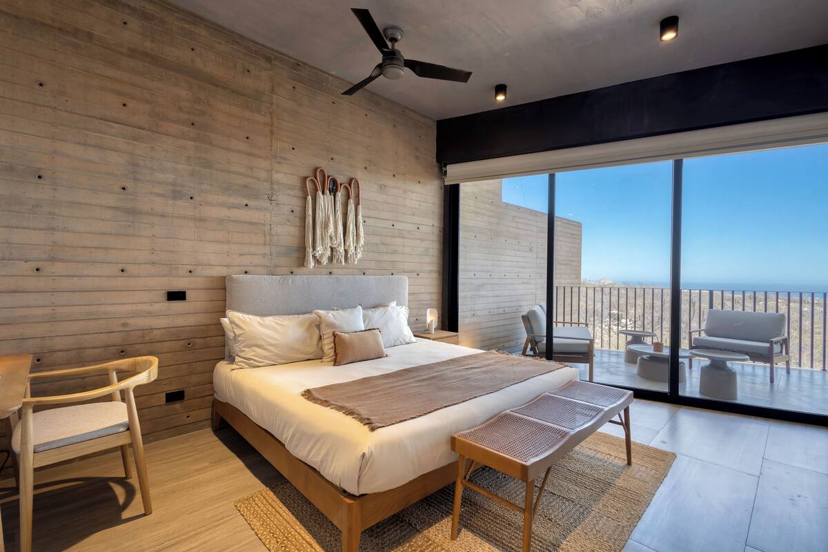 A modern bedroom with a large bed, a ceiling fan, and a view of the beach through large windows.