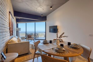 A modern dining area with a wooden table set for four, a sofa, and a view of the ocean through large windows. - OR Cabo Boutique Residences by Lumina (Cabo San Lucas)