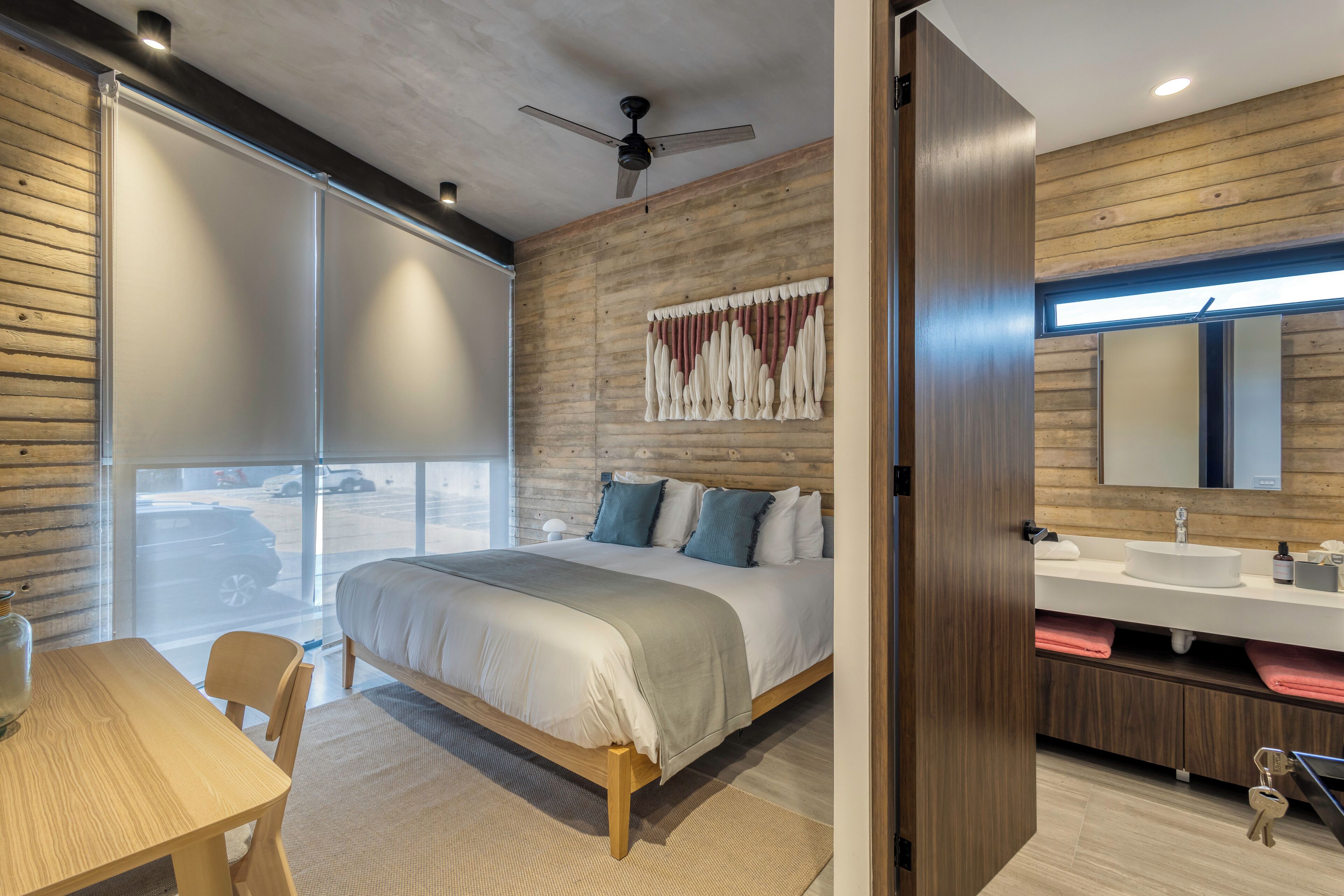 Ein modernes Schlafzimmer mit einem großen Bett, einem Holz-Kopfteil, einem Deckenventilator, einem Badezimmer mit Waschbecken und Spiegel sowie Blick ins Freie.
