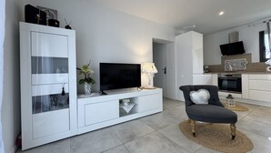 A modern living room with a flat-screen TV, a white entertainment unit, a grey armchair, and a kitchen area with a TV and oven. - House in Ruaudin Near Le Mans Circuit (Ruaudin)