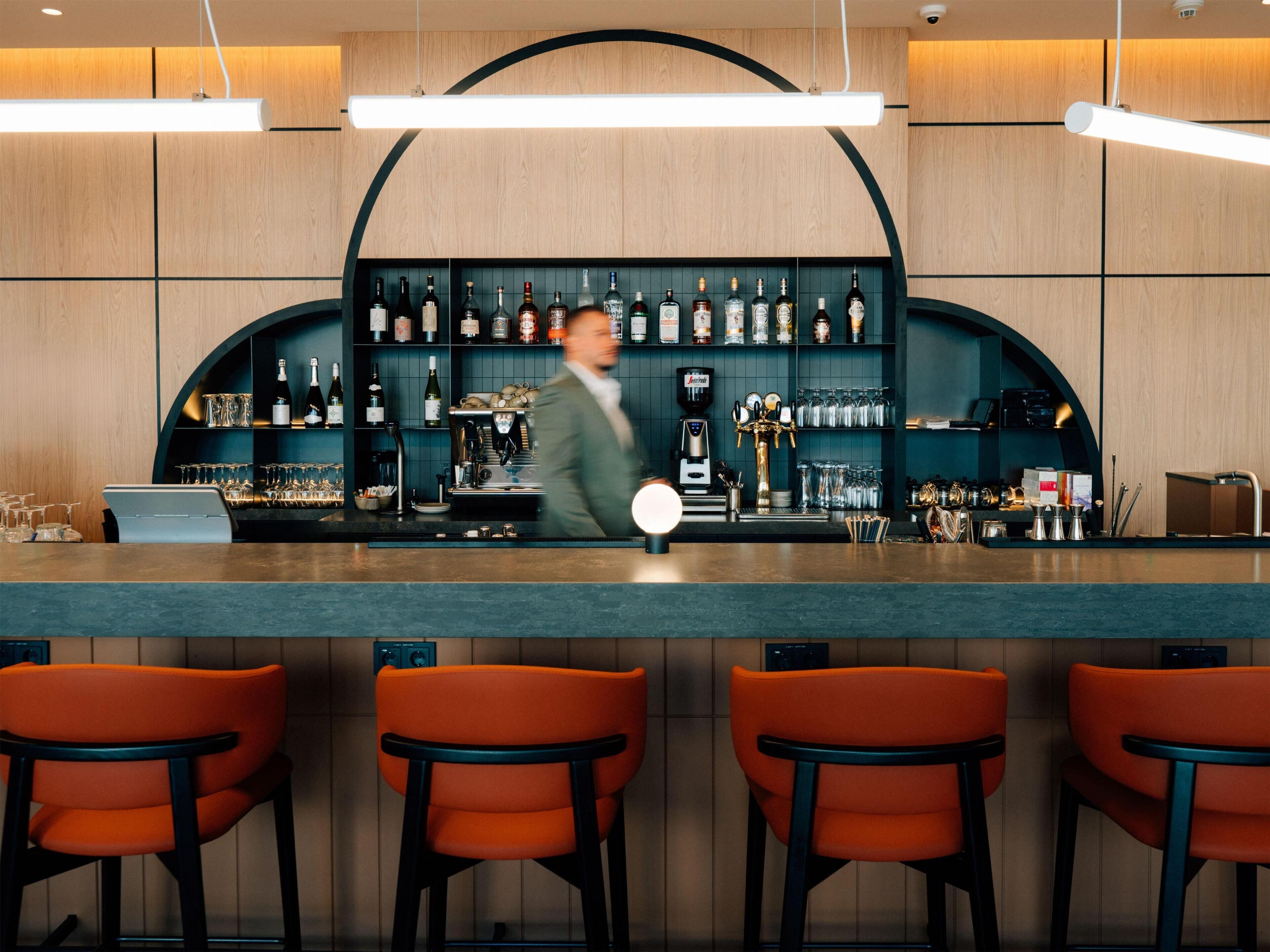 A bar area with a counter, shelves of bottles, and orange chairs.