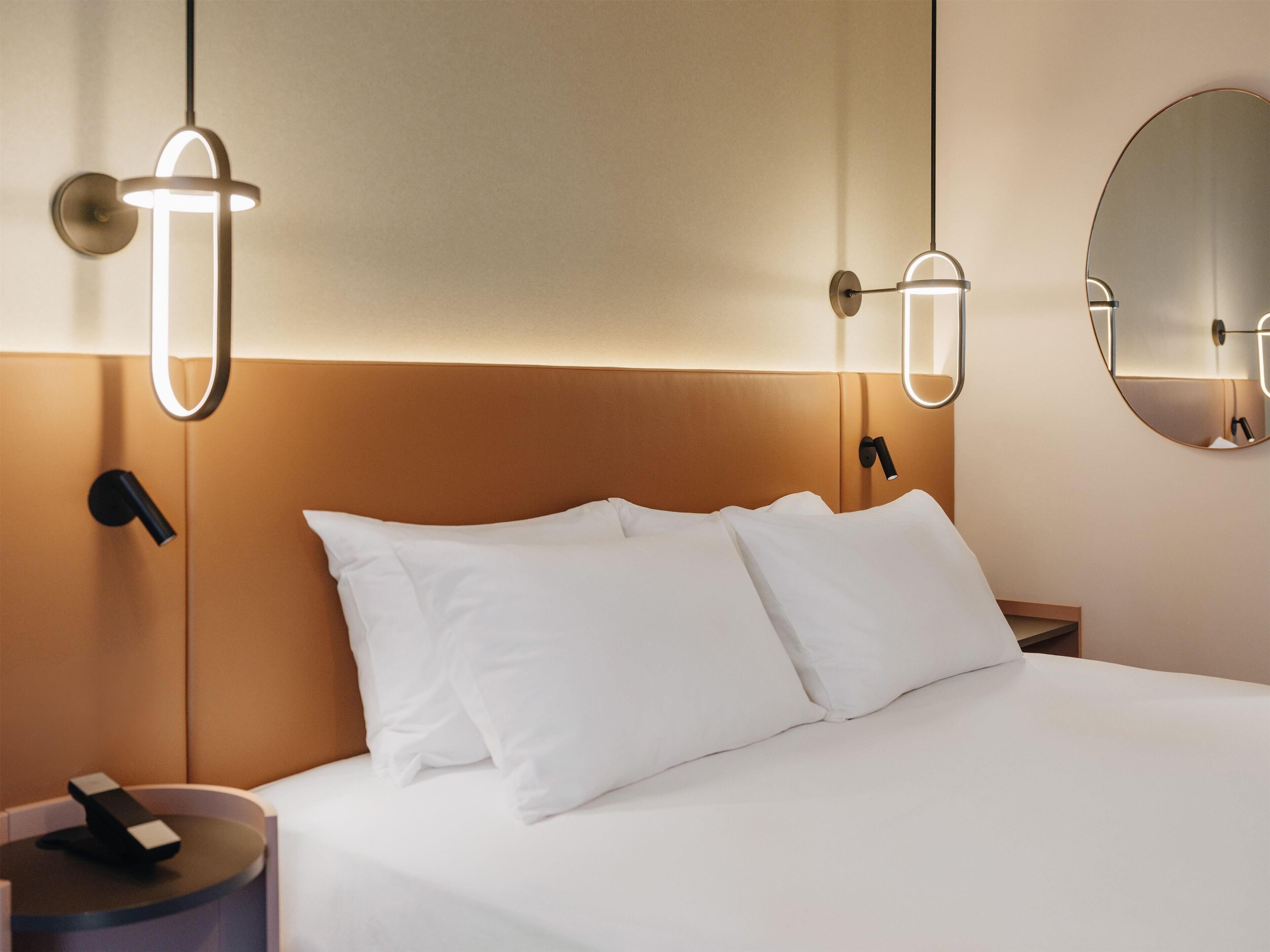 A hotel room with a bed, bedside tables, a round mirror, and wall-mounted lighting fixtures.