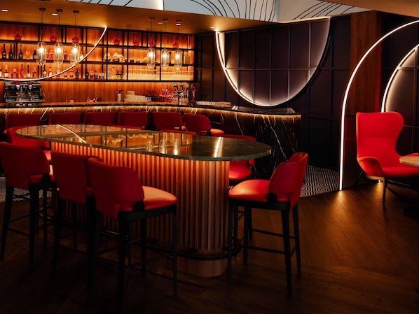 A bar area with a round table surrounded by red chairs, a wooden bar counter with bottles and glasses, and a modern ceiling design. - Hyatt Place Tallinn (Tallinn)
