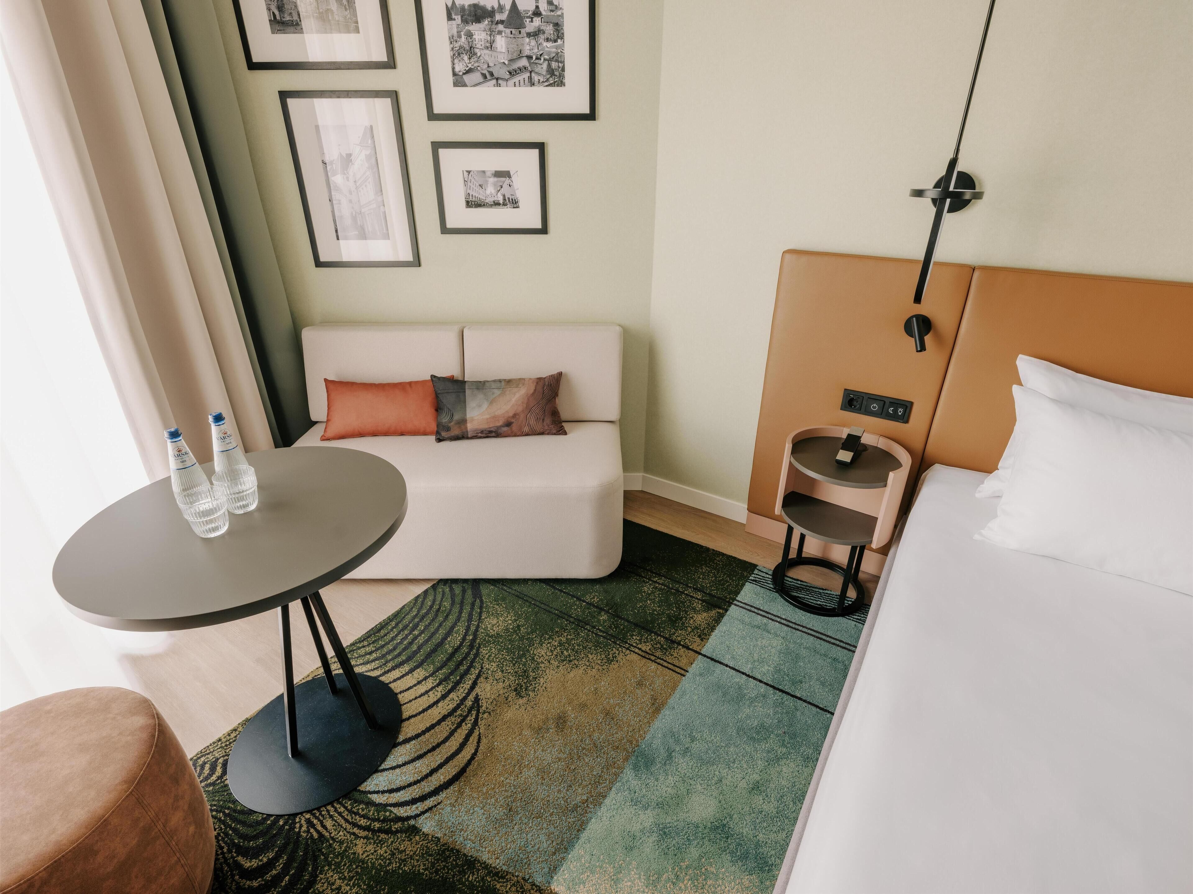 Ein modernes Hotelzimmer mit einer Couch, einem kleinen runden Tisch mit zwei Flaschen, einem Bett, einem Nachttisch und einer Wandlampe.
