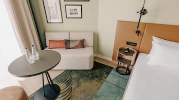 Una habitación de hotel moderna con un sofá, una mesita redonda pequeña con dos botellas, una cama, una mesita de noche y una lámpara de pared.