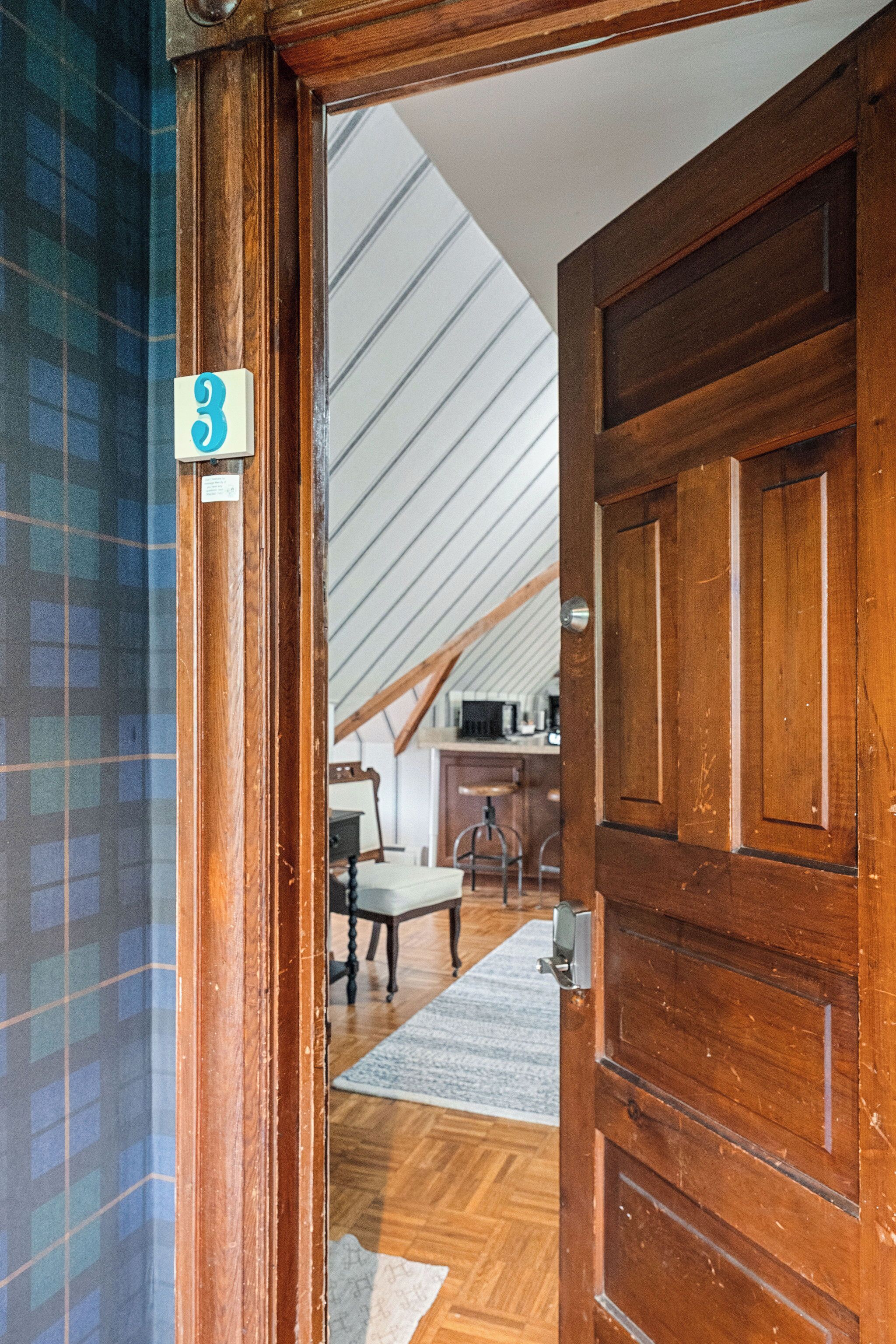 A wooden door with a number plate, a room with a sloped ceiling, and a view of a kitchen area.