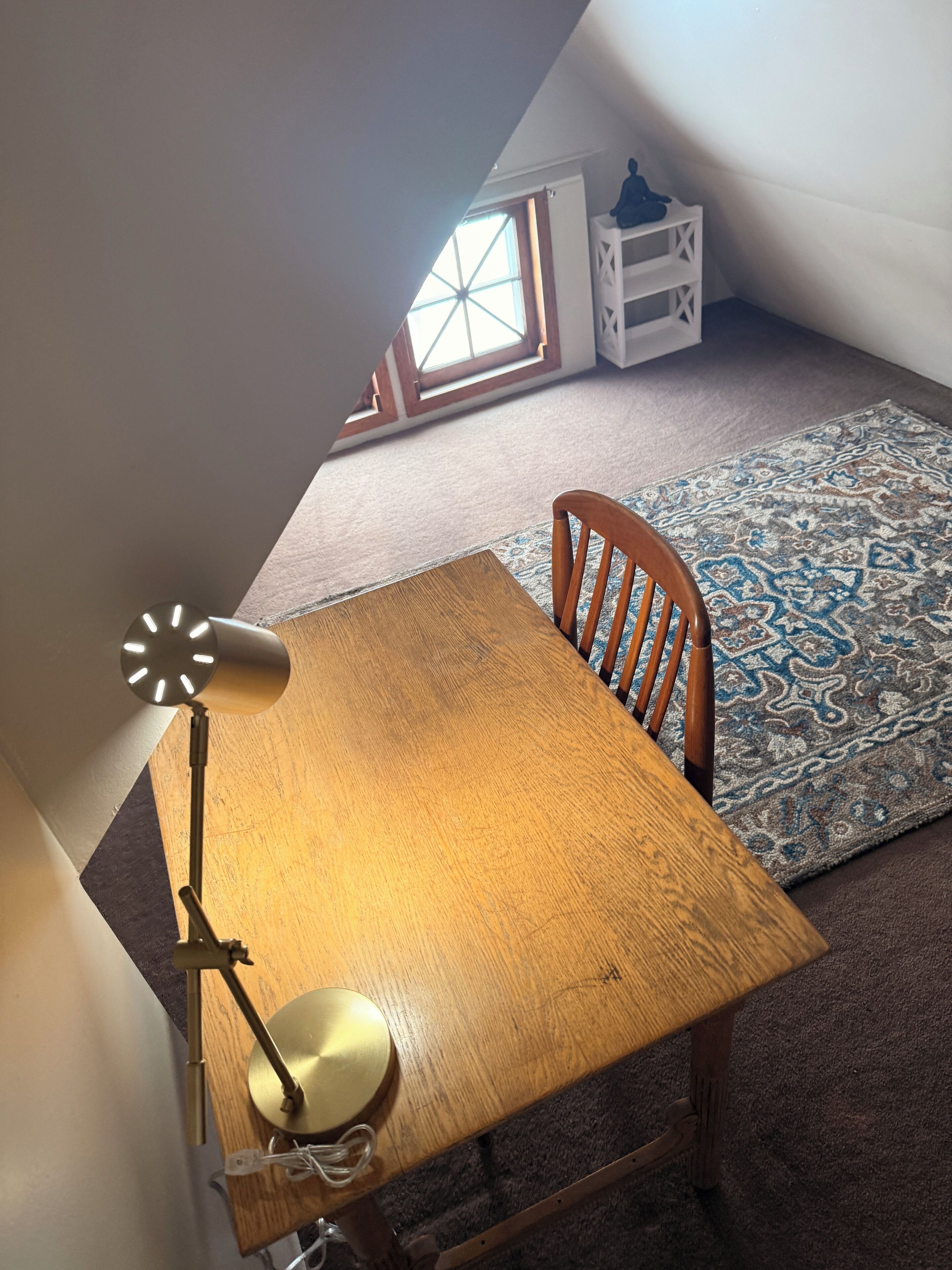Una habitación con una mesa y silla de madera, una lámpara, un tapete estampado y un ventanal con vistas al exterior.