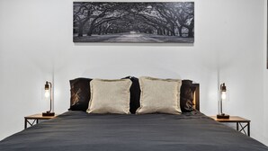 Une chambre à coucher avec un grand lit, deux tables de chevet et un tableau représentant une route bordée d’arbres, accroché au mur.