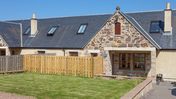 Une petite maison en pierre avec une clôture en bois, une cour gazonnée et un chemin pavé menant à l’entrée.