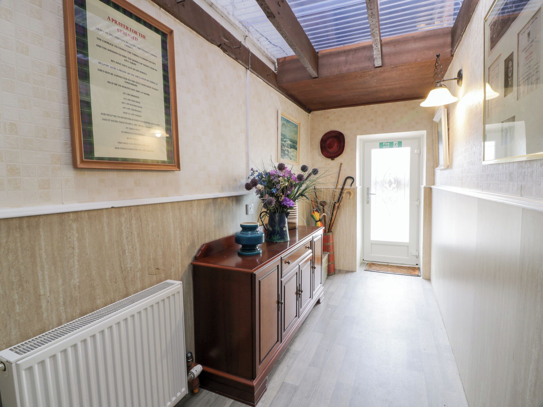 A hallway with a framed prayer for the sick, a radiator, a wooden cabinet, and a door with a fire exit sign.