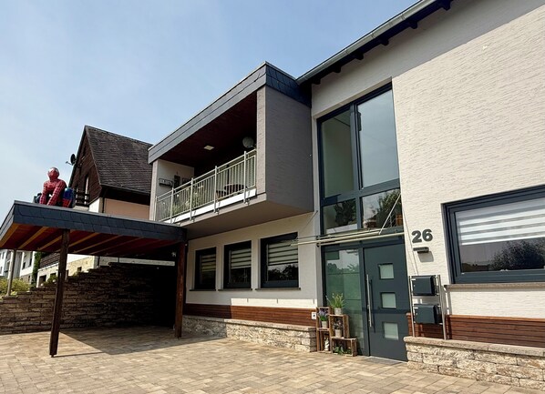 Una moderna casa a due piani con un balcone, ampie finestre e un portone garage con il numero 26.