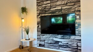 Una sala de estar con una pared de piedra, un televisor de pantalla plana, una planta colgante y una mesita con una lámpara.